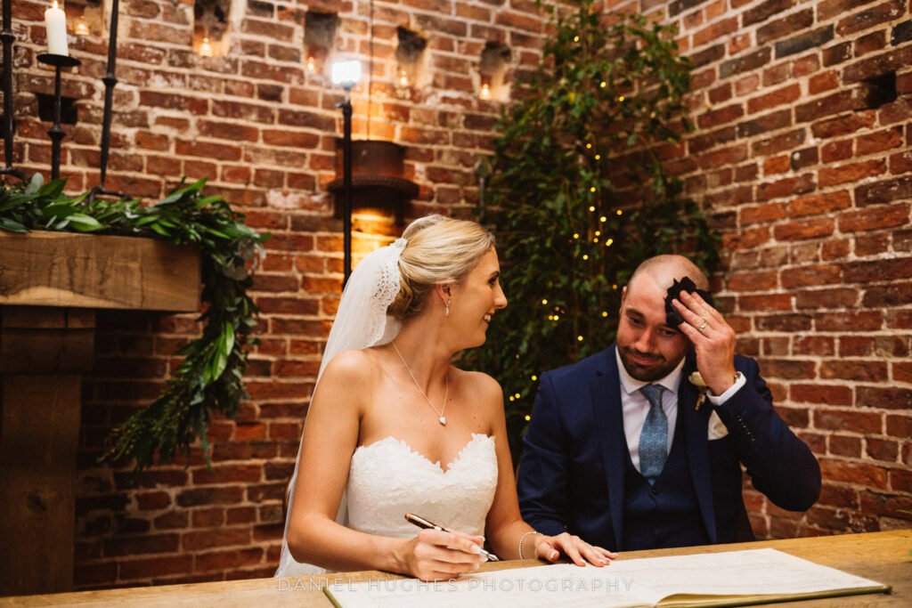 Bride and groom sitting down to sign their register, the groom is wiping his forehead with a napkin as the bride smiles fondly at him.