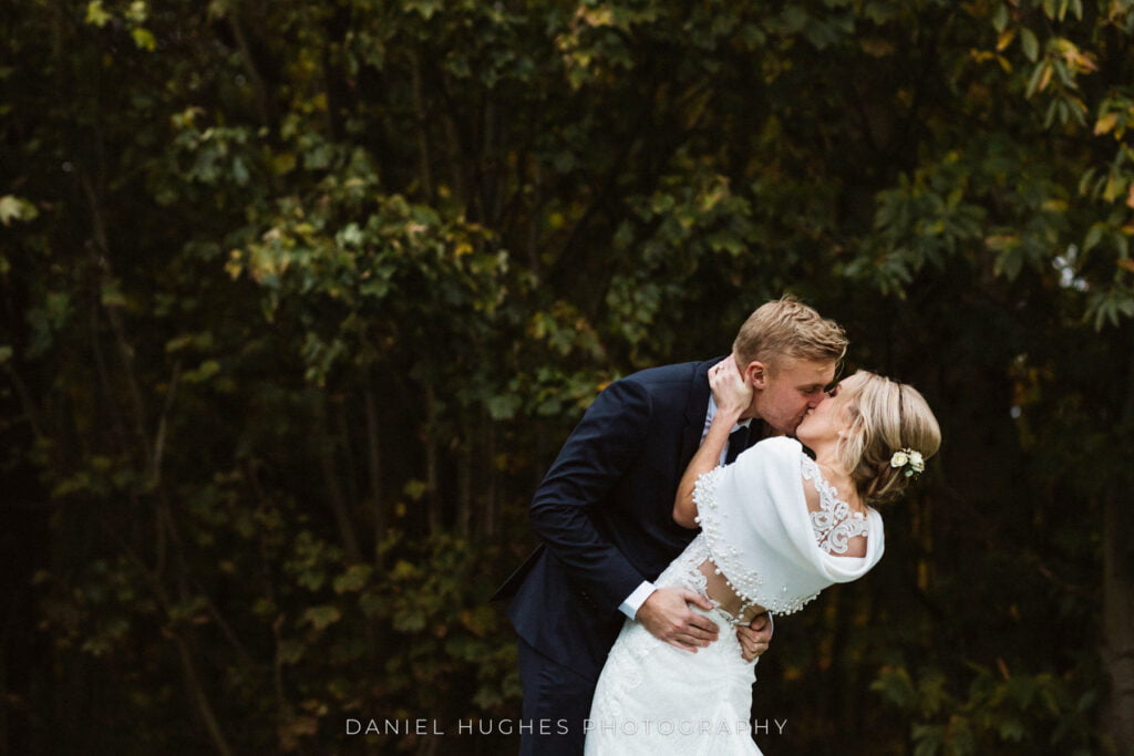A stunning photograph of the groom holding the bride as they kiss. The bride leans backwards, framed by the blurry trees in the background.