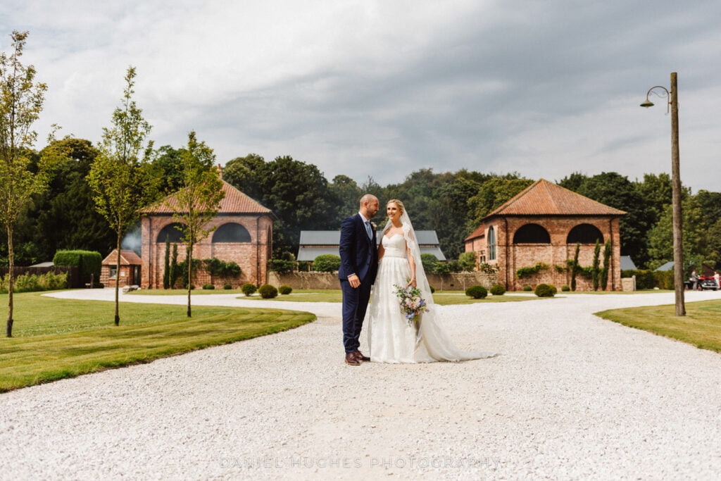 A couple smiles standing on the driveway in front of Hazel Gap Barn with the towers behind them
