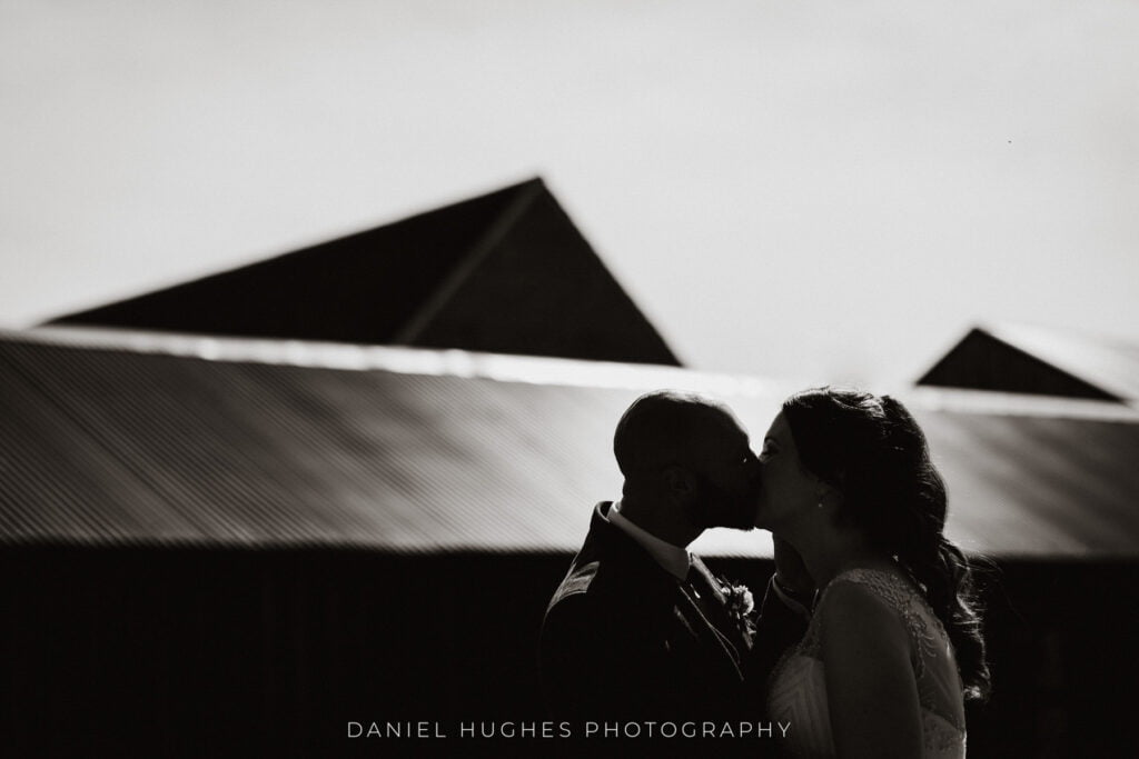 A stunning black and white close-up of a couple kissing, positioned in the bottom half of the frame to the right. They are partially silhouetted against the bright sunlight reflecting off the metal roof. 