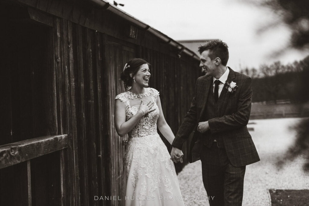 A couple walks through the grounds at Hazel Gap, smiling at each other. The bride holds one hand to her chest and his in the other. The image is in black and white.