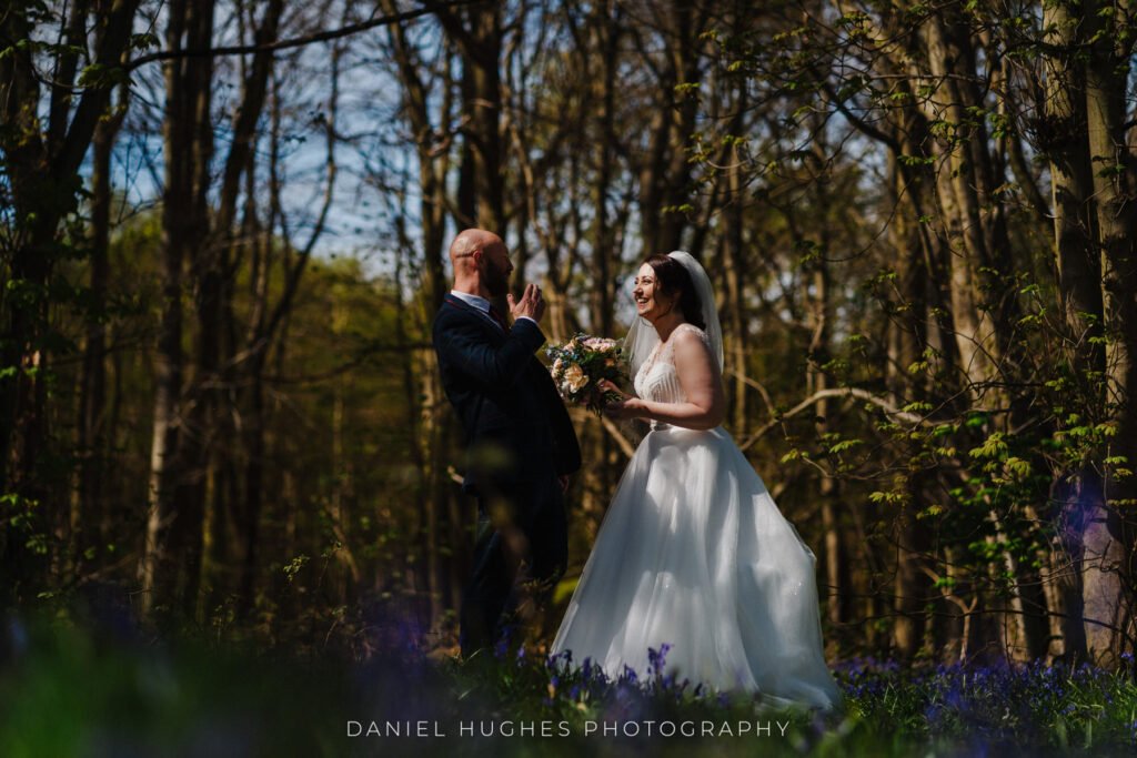 Shot of the bride and groom laughing in the woods at the back of Hazel Gap. Sunlight streams through the trees, illuminating the bride perfectly, with bluebells barely visible covering the floor.