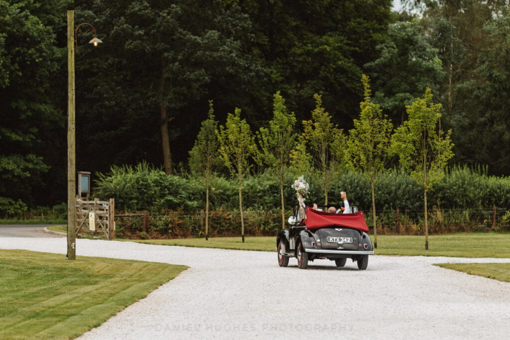 A couple waving from the back of a vintage car as they drive down the tree-lined driveway at Hazel Gap