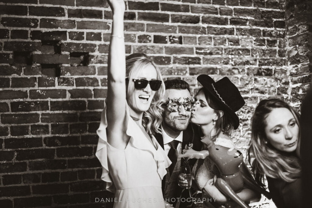 A black and white image of guests smiling with sunglasses on while operating the Flash Bstrds photo booth in the main barn. They are using the brick wall as their backdrop.