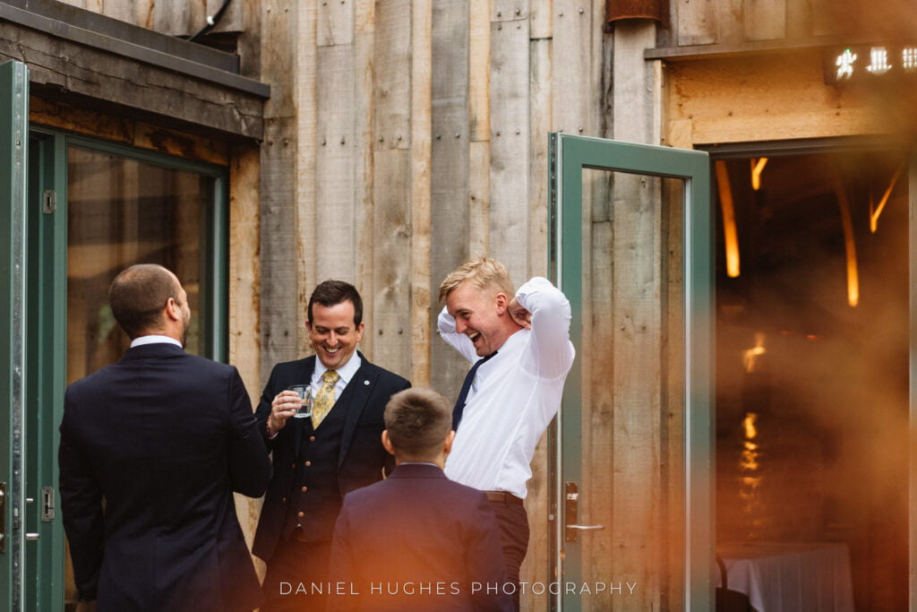 Guests standing outside with drinks, the groom is throwing his head back laughing as he attempts to put on his tie.
