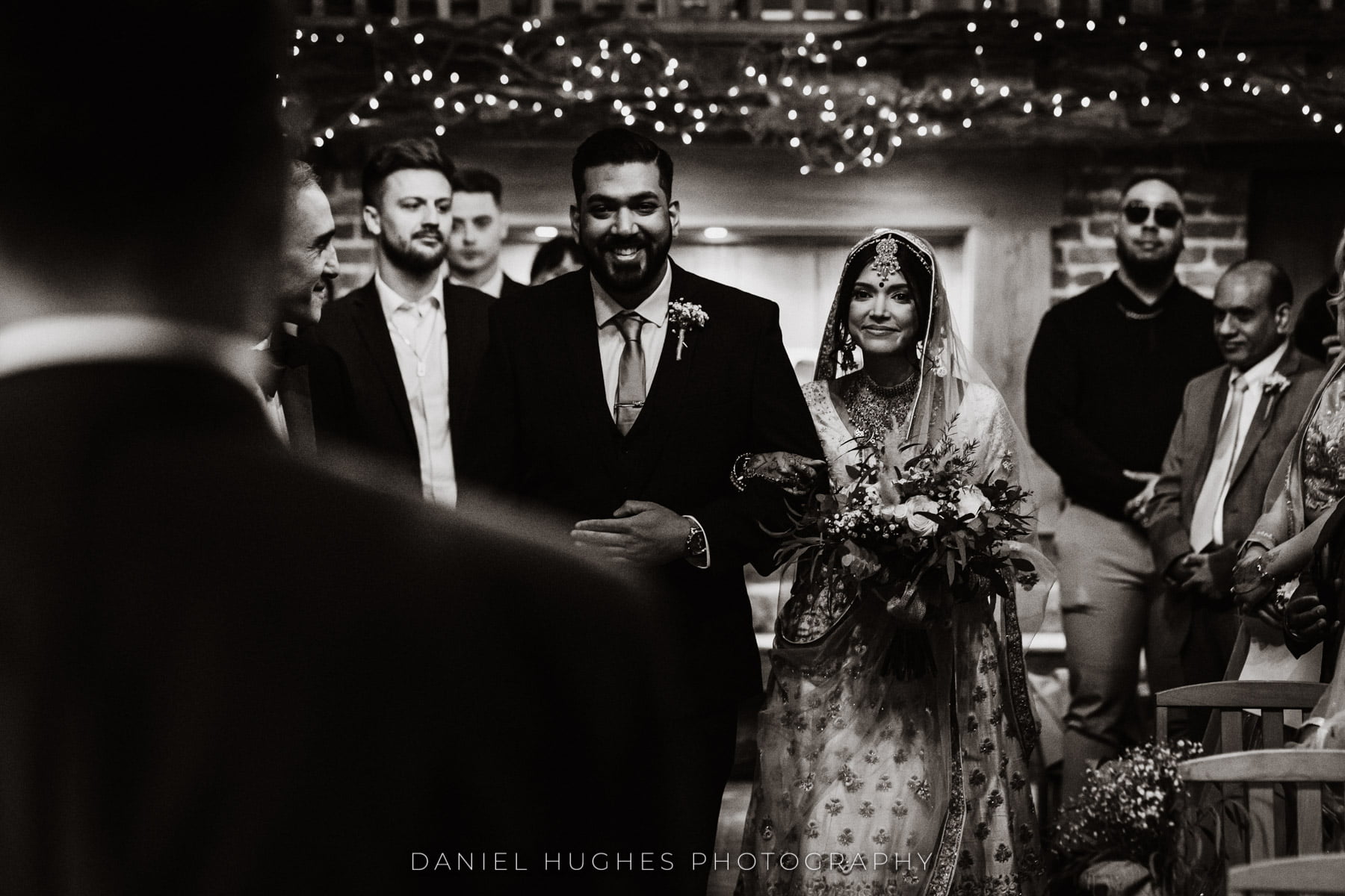 Black and white photograph, shot over Gurpreet's shoulder. An excited Kamrun is being walked up the aisle by her smiling brother.